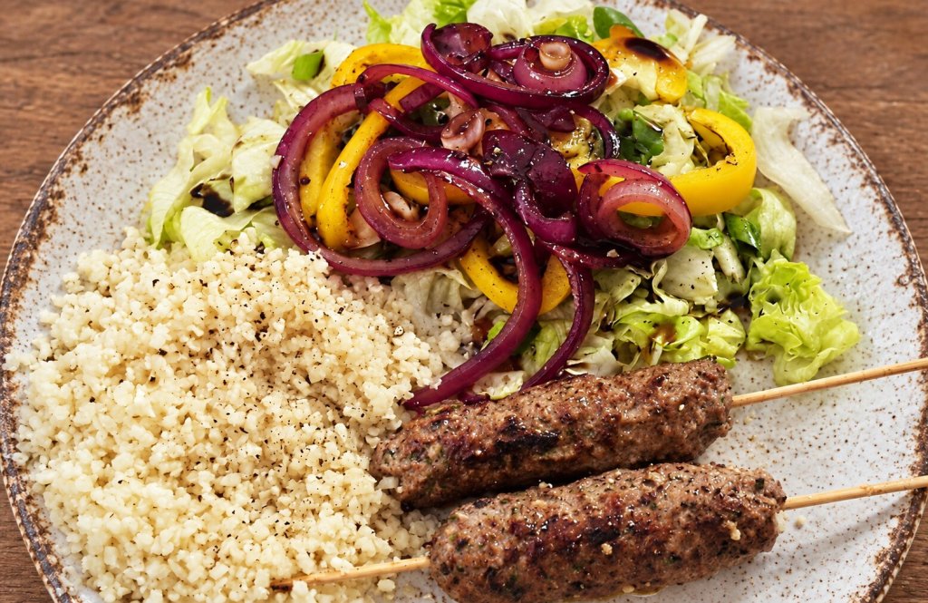 Minted lamb kebabs with balsamic onions, cauliflower rice and salad on a rustic off-white ceramic plate