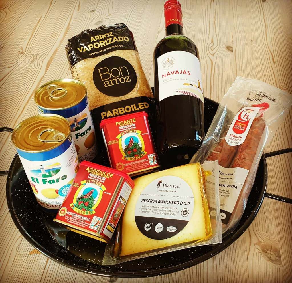 Spanish pantry ingredients arranged in a paella pan, including rice, paprika, chorizo, Manchego and wine